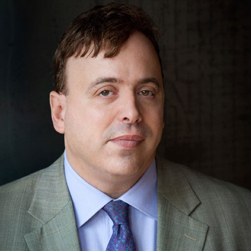 The image shows a man with glasses, wearing a suit, standing against a dark background.