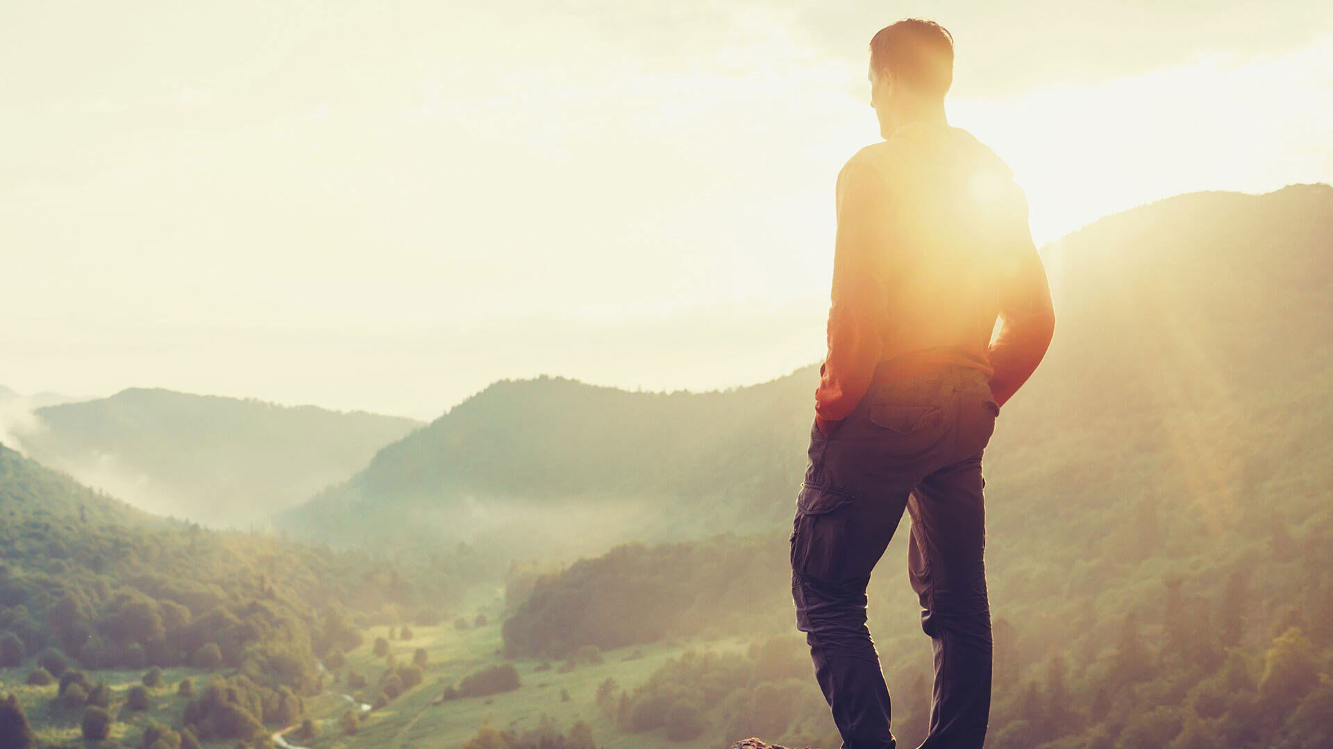 A man stands on a hilltop overlooking a valley under a sunset sky, with mountains in the background.