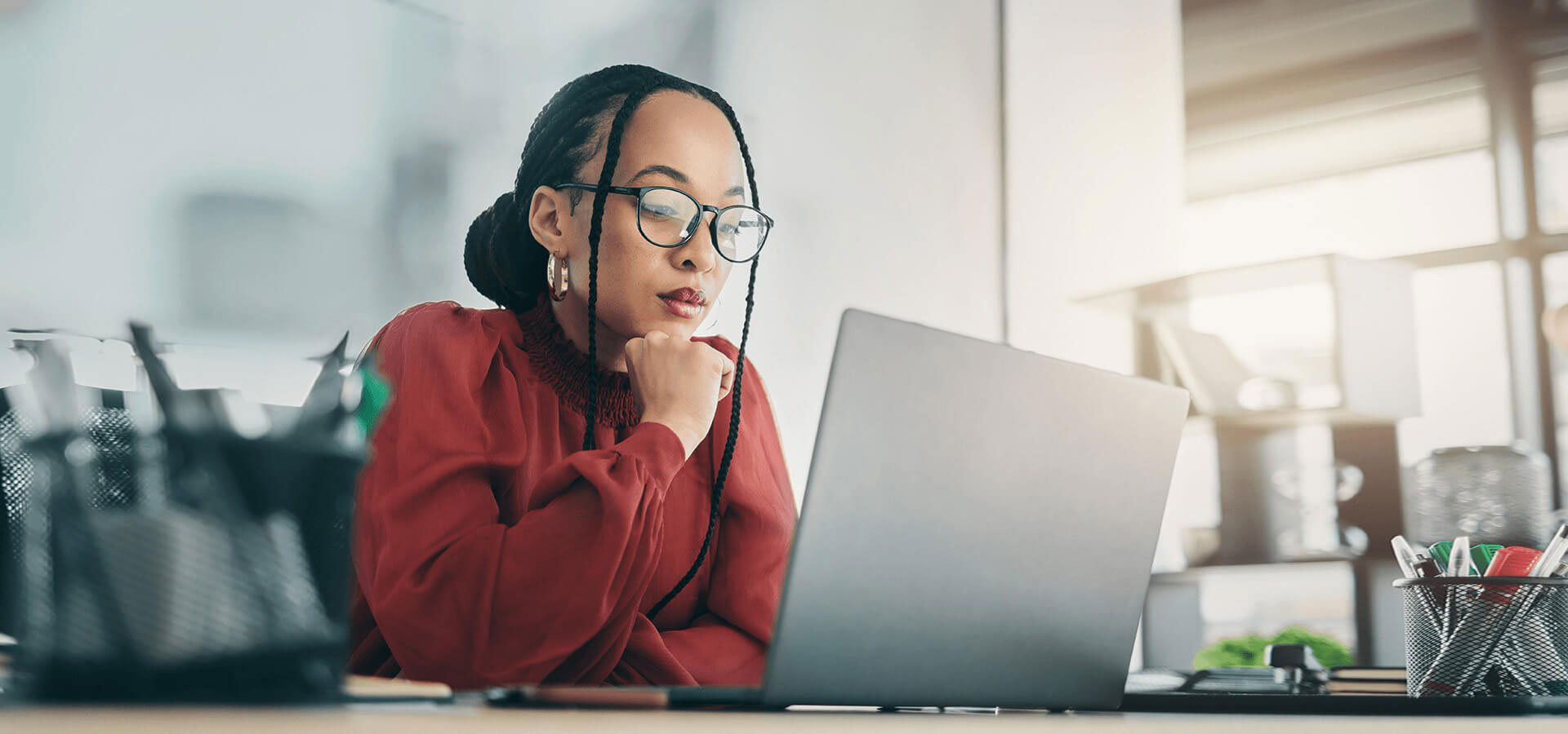 A woman wearing glasses is seated at a desk with a laptop, working intently in an office setting.